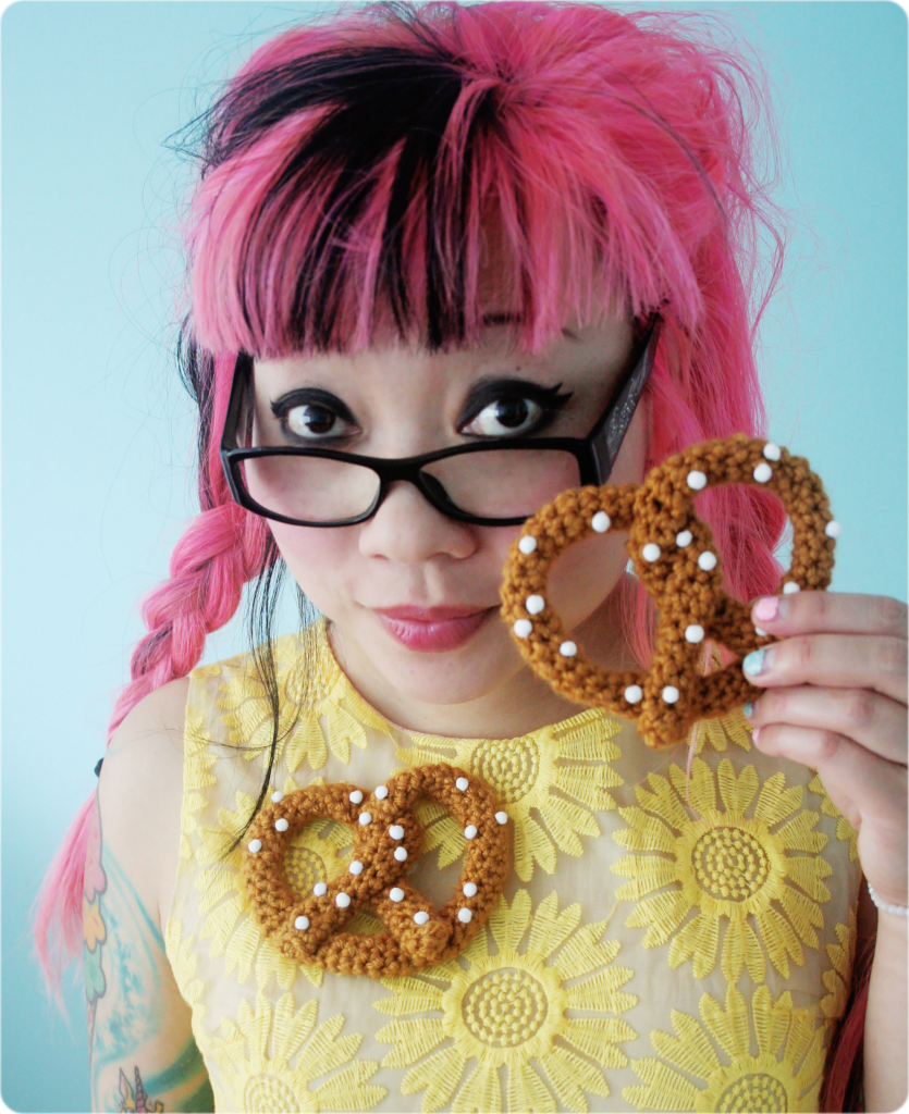 Twinkie Chan wears a yellow floral dress and a crocheted pretzel brooch. She is also holing a crocheted pretzel in her hand.