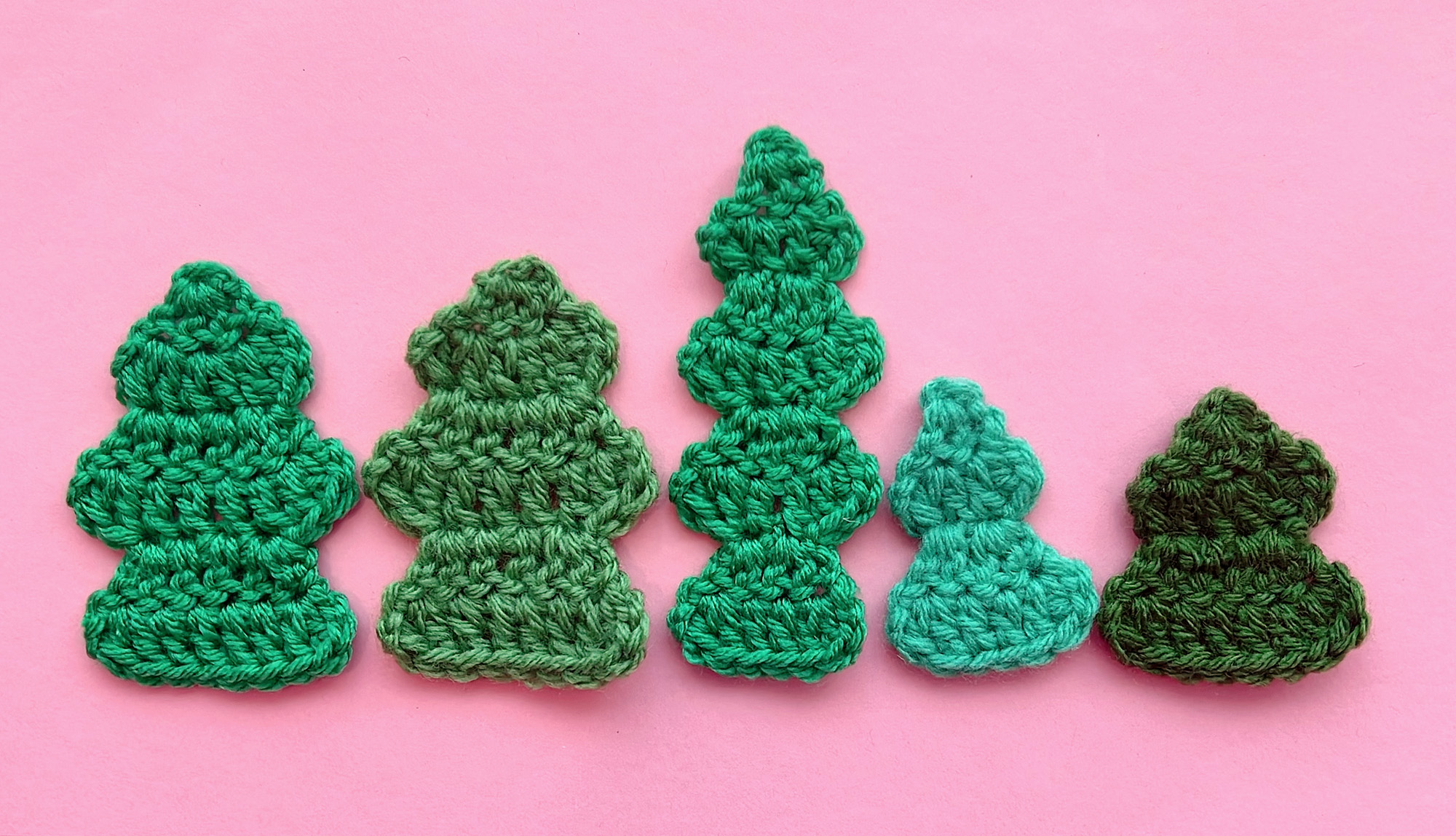 5 small crocheted Christmas tree appliqués in a horizontal row in various shades of green and different heights on a pink background