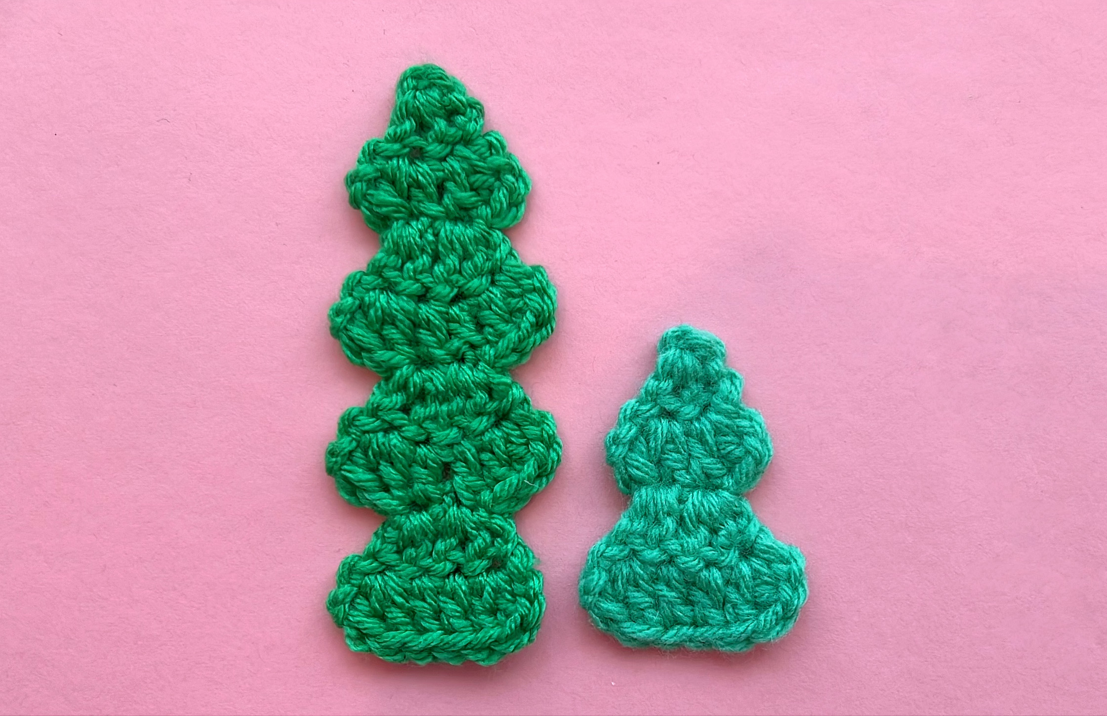 2 small flat crocheted Christmas trees of different heights and different shades of green on a pink background